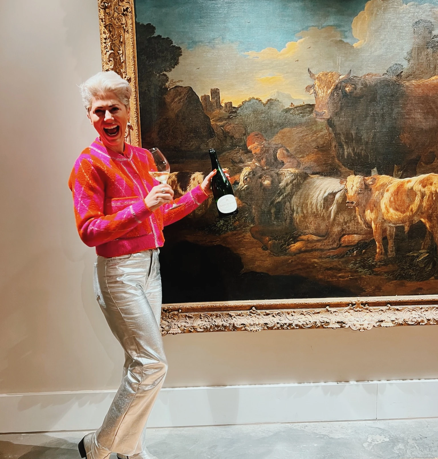 Person holding a wine glass and bottle beside a large framed painting of cattle