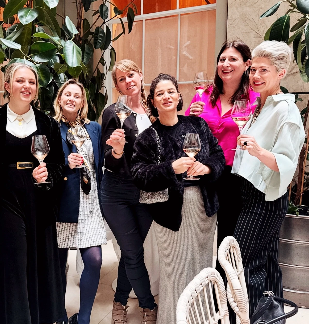 Six women pose together indoors, holding glasses of rosé wine among large plants.