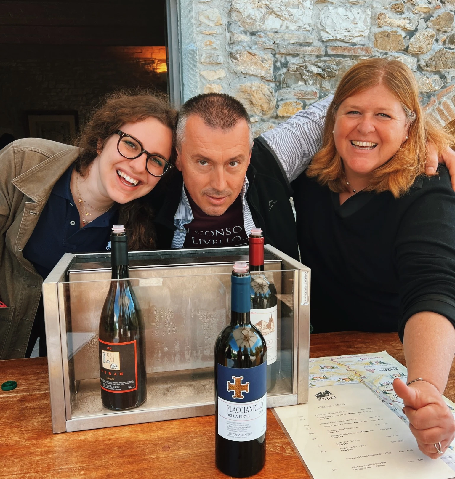 Three people pose behind two wine bottles and a tasting menu at a Tuscan winery