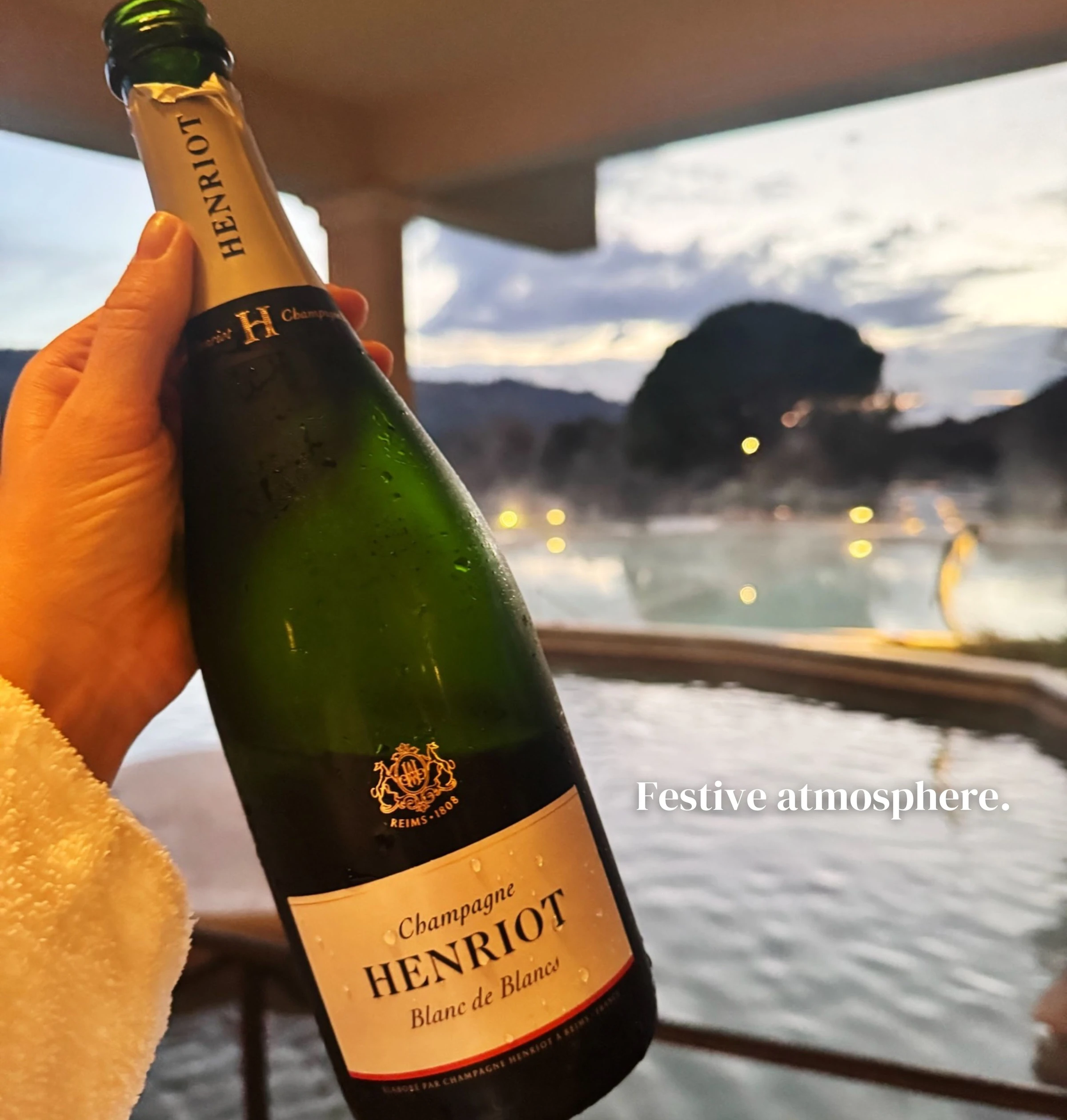 Hand holding a bottle of Henriot Blanc de Blancs Champagne beside an outdoor spa pool