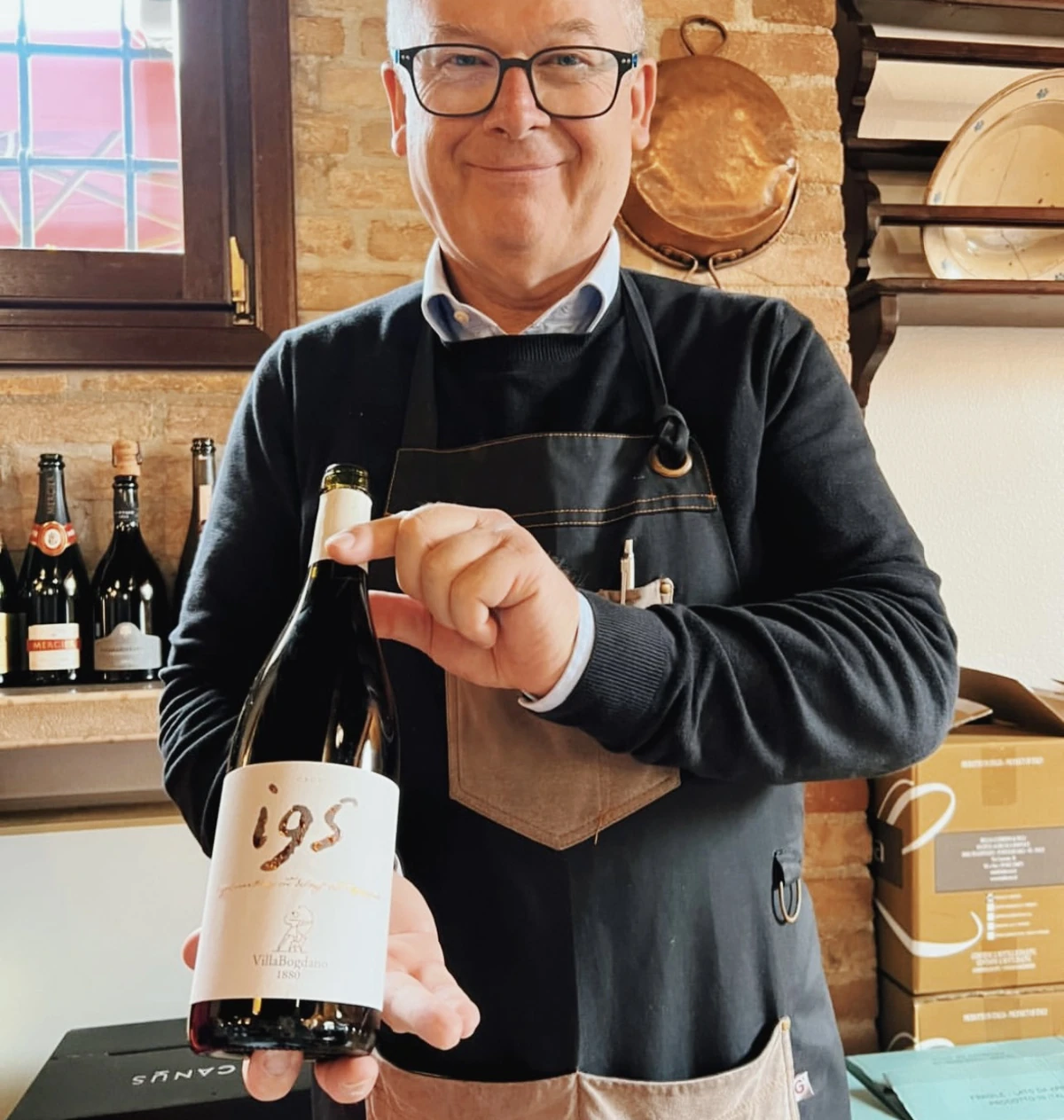 Domenico Veronese holds a bottle of 195 wine at a winery, with wine bottles and barrels behind him.