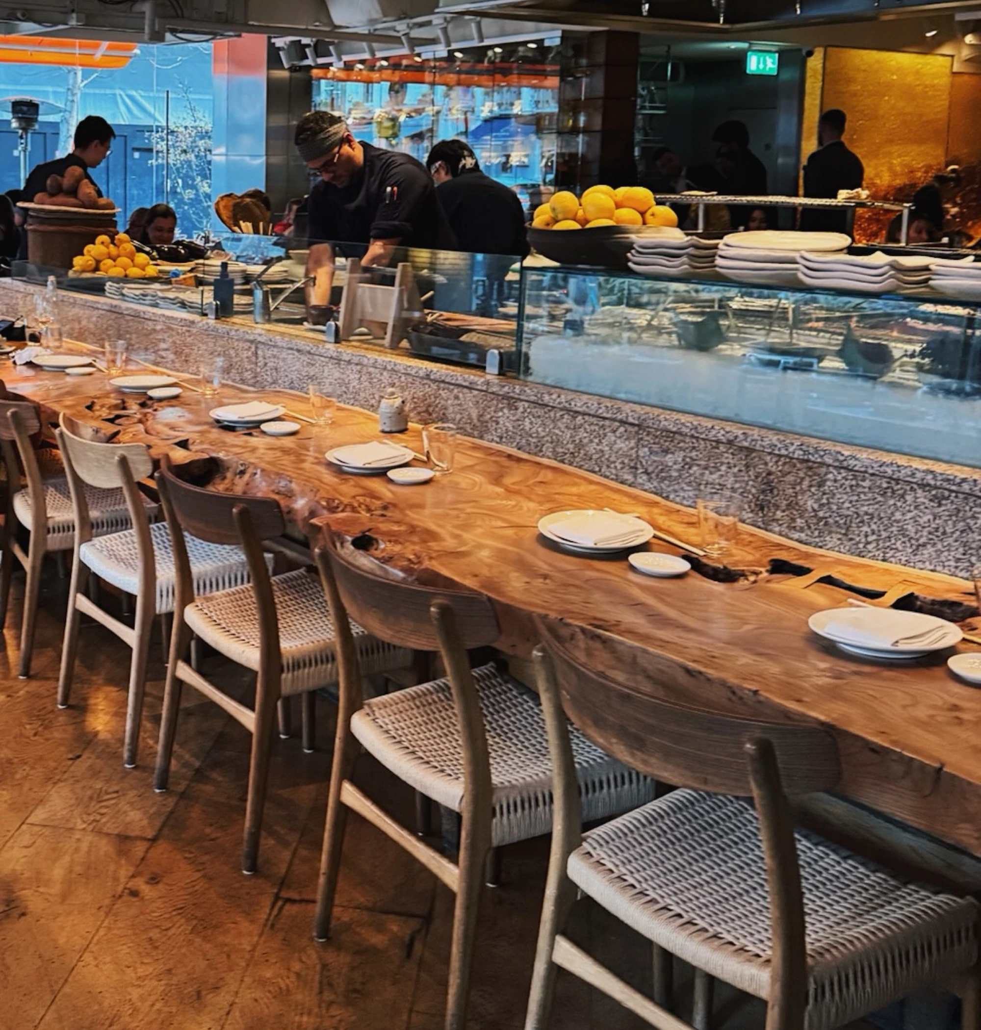 Restaurant counter with chefs preparing food and a long table set with plates and glasses