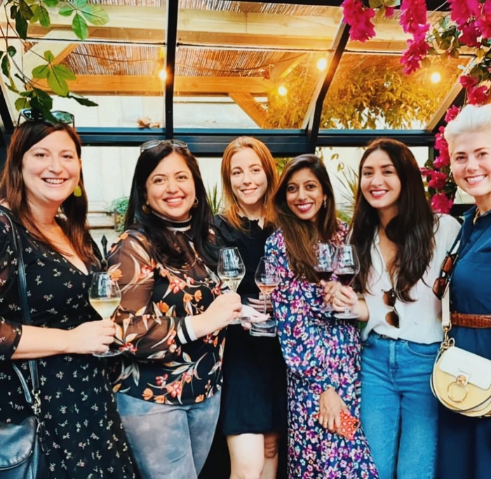 Six women smile for a group photo while holding wine glasses in a bright, flower-filled venue.