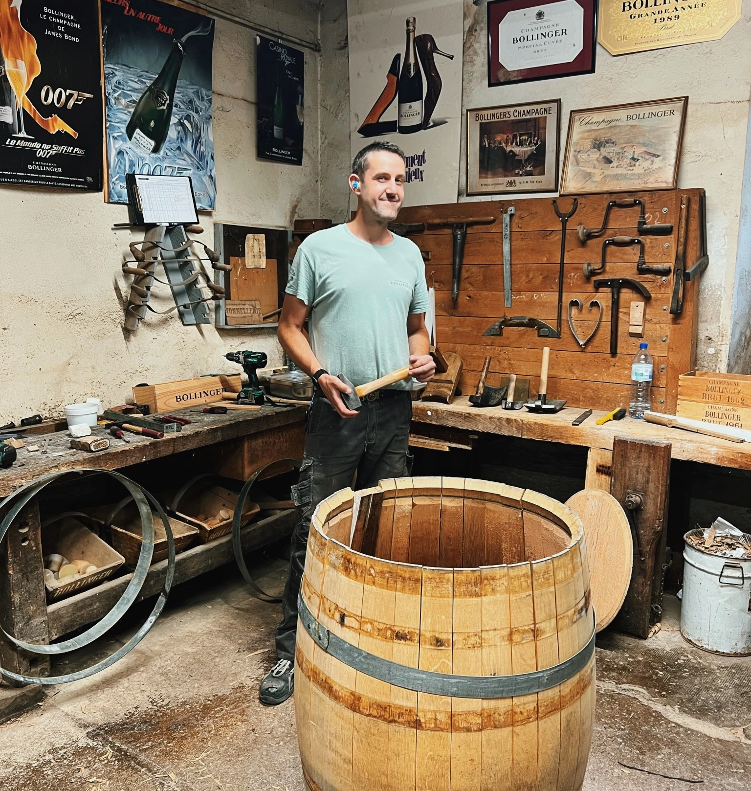 Man standing beside an open oak barrel in a workshop decorated with Bollinger Champagne posters and tools