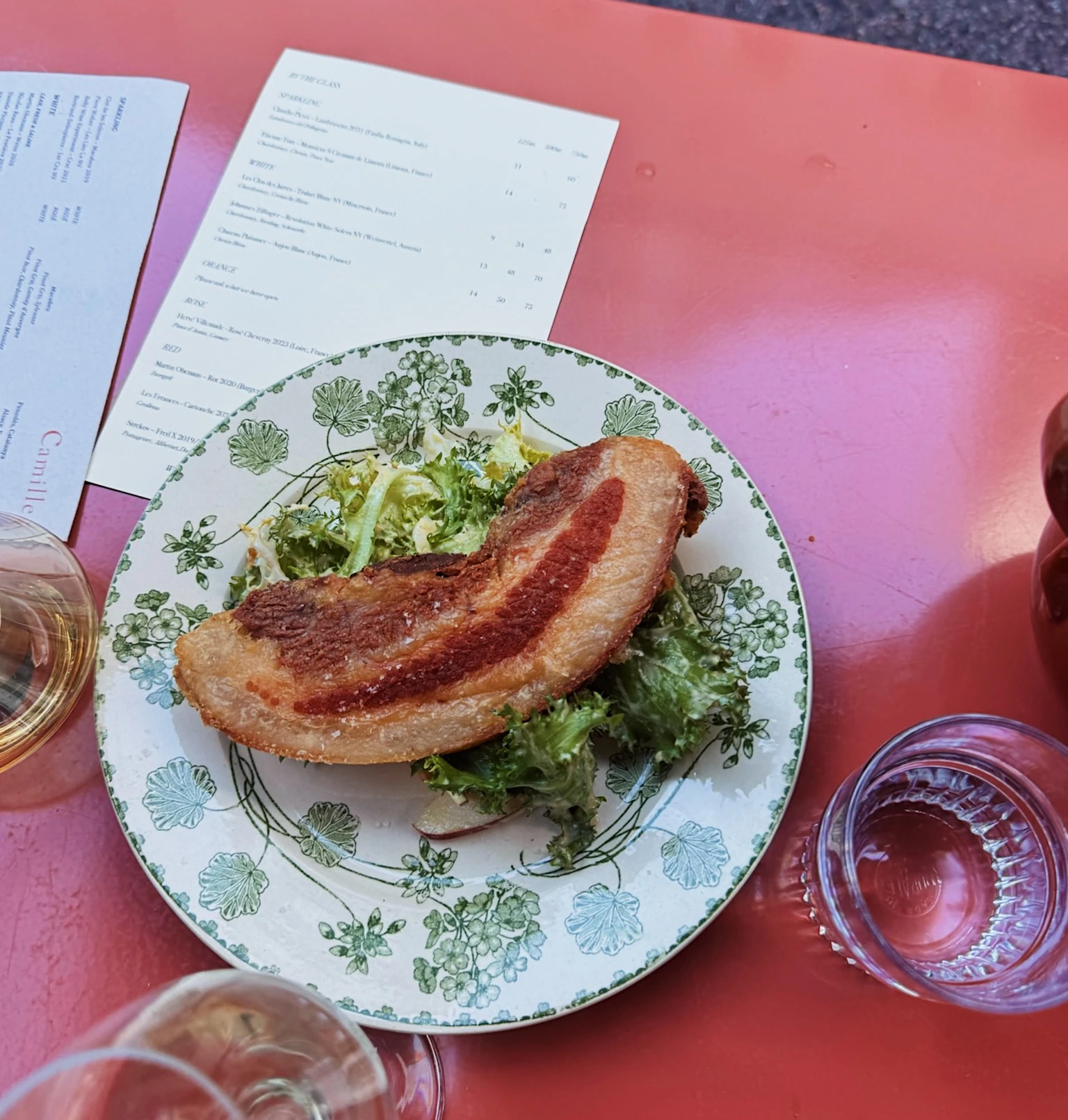 Crisp pork belly with salad on a patterned plate, surrounded by wine glasses and menus