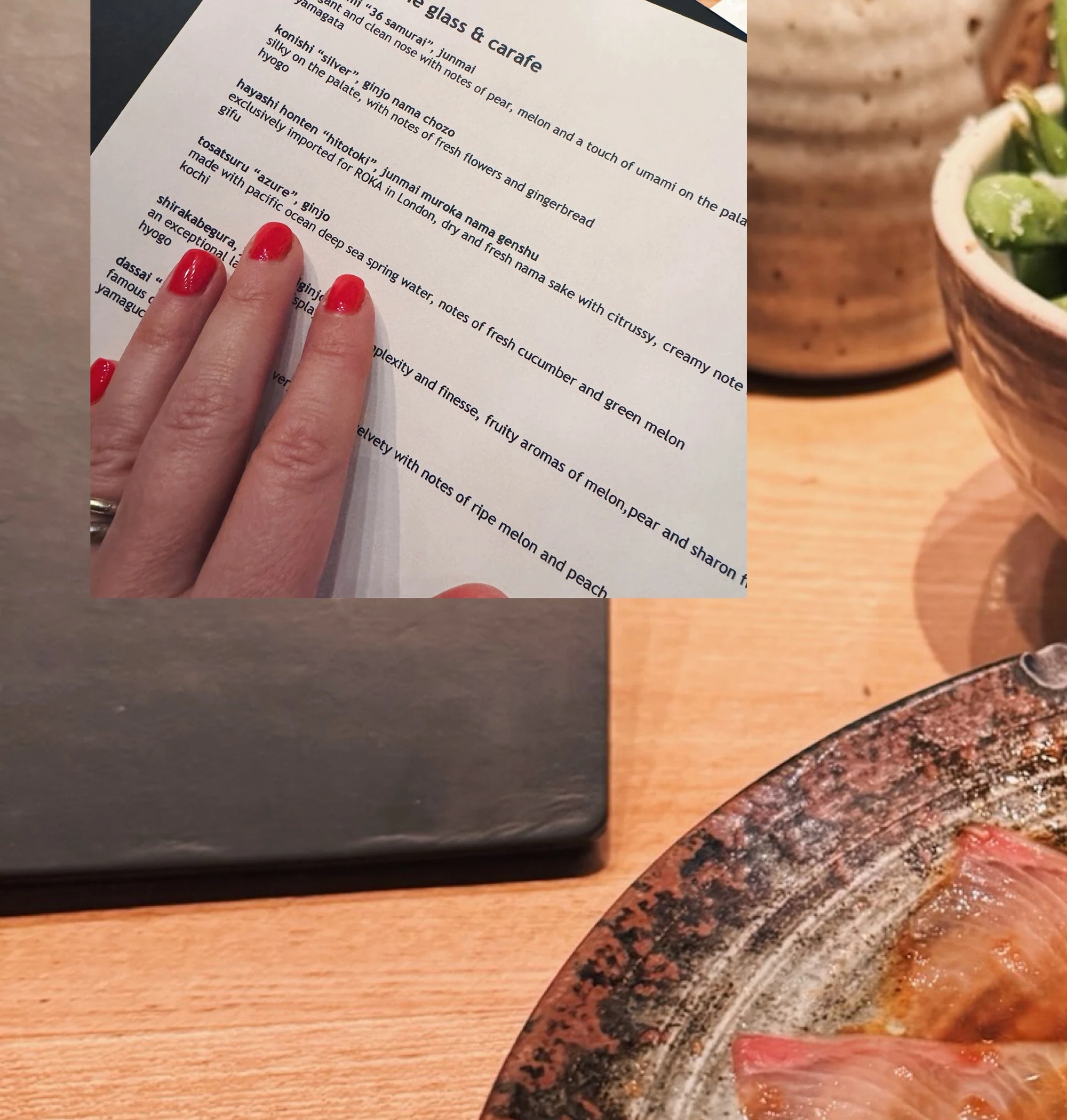 Sake tasting menu beside sushi and a hand with red-painted nails