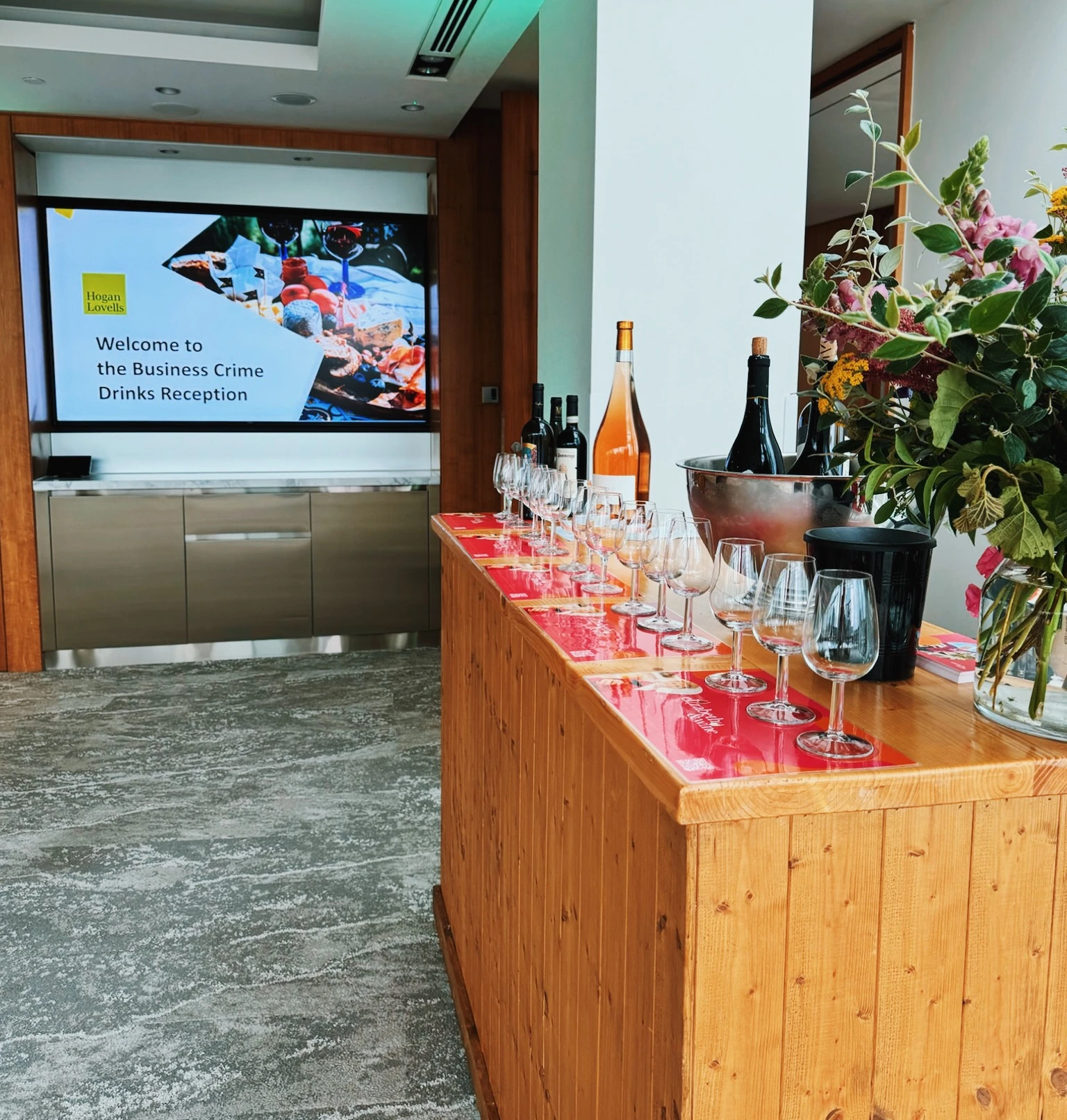 Wine tasting table with glasses, bottles and flowers beside a screen welcoming guests to a business drinks reception