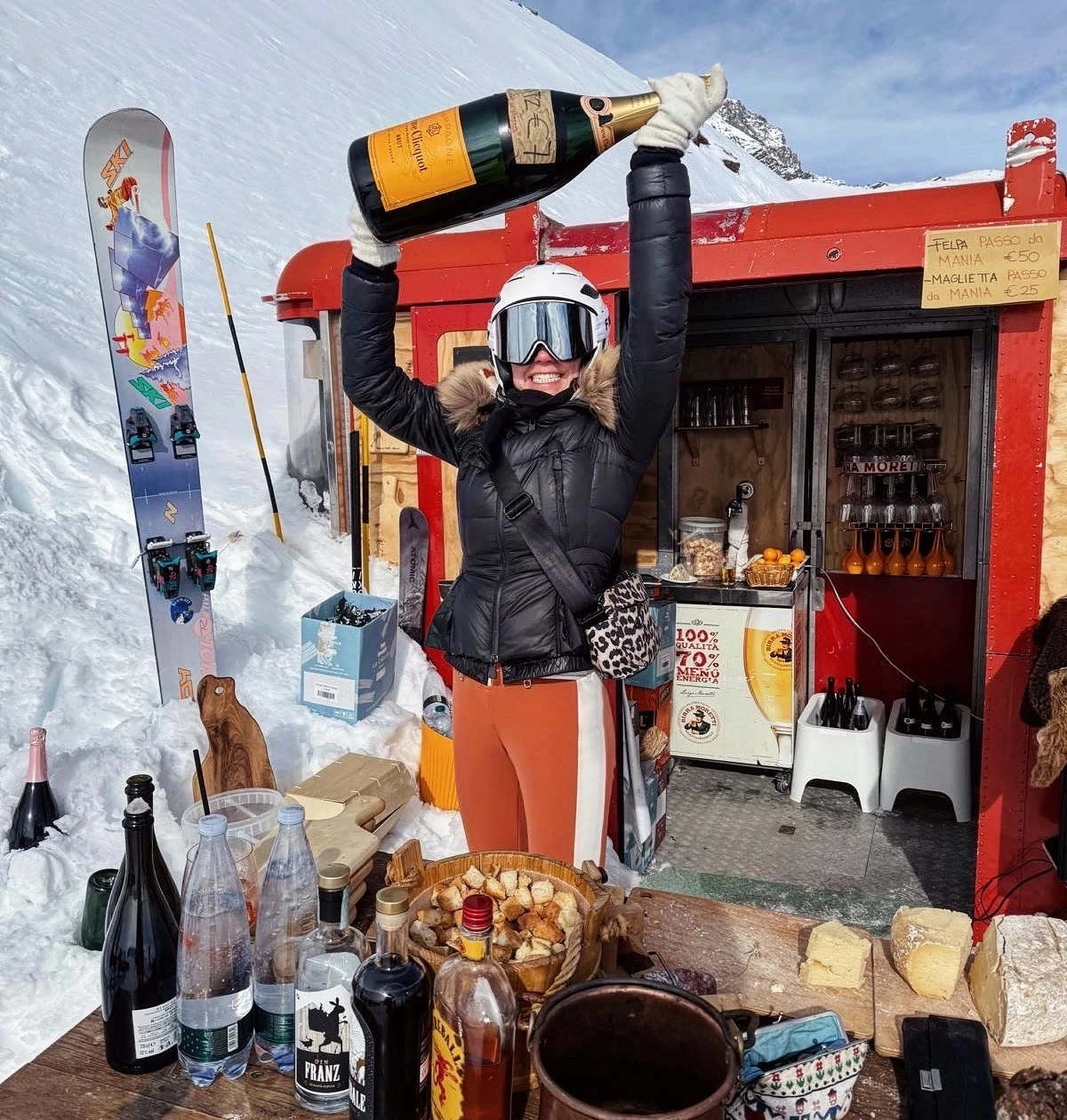 Skier raising a bottle of Champagne at a snowy mountain bar, with skis, drinks and food on the counter