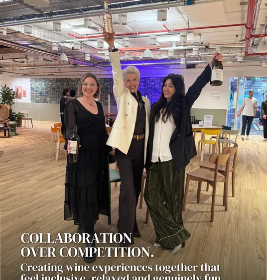 Three women smiling and raising wine bottles in a bright event space