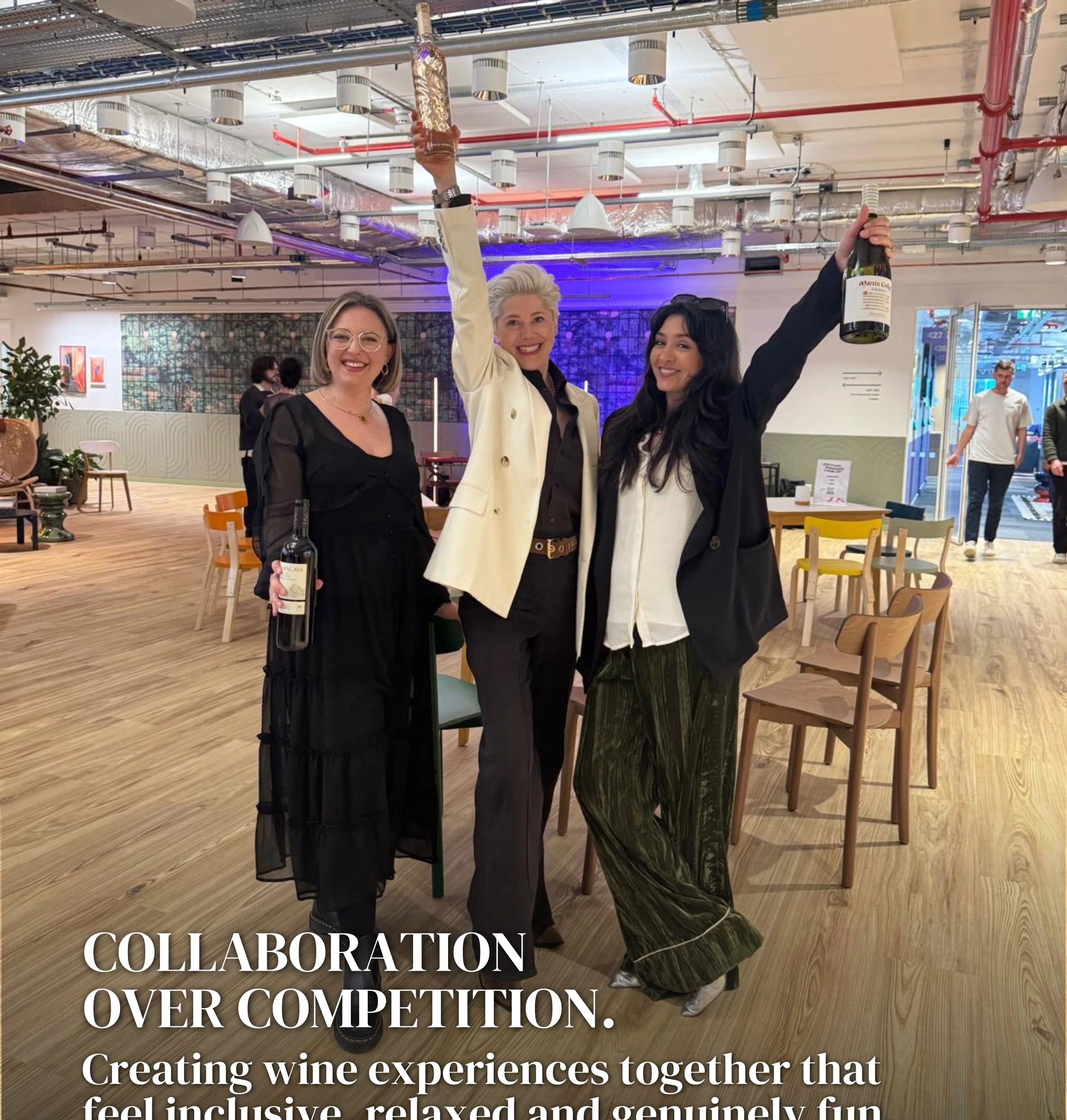Three women smiling and raising wine bottles in a bright event space