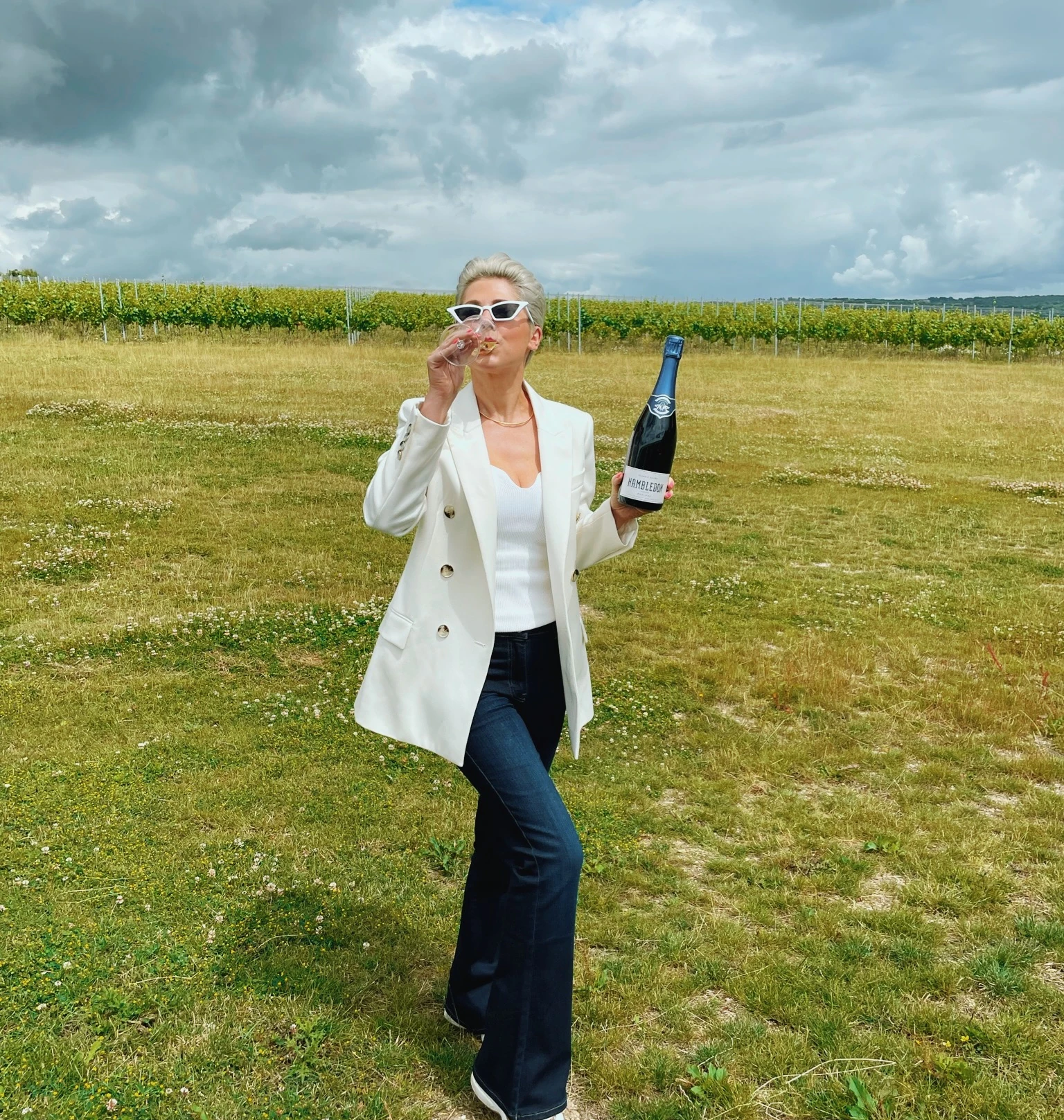 Person drinking sparkling wine beside rows of vines under a cloudy sky