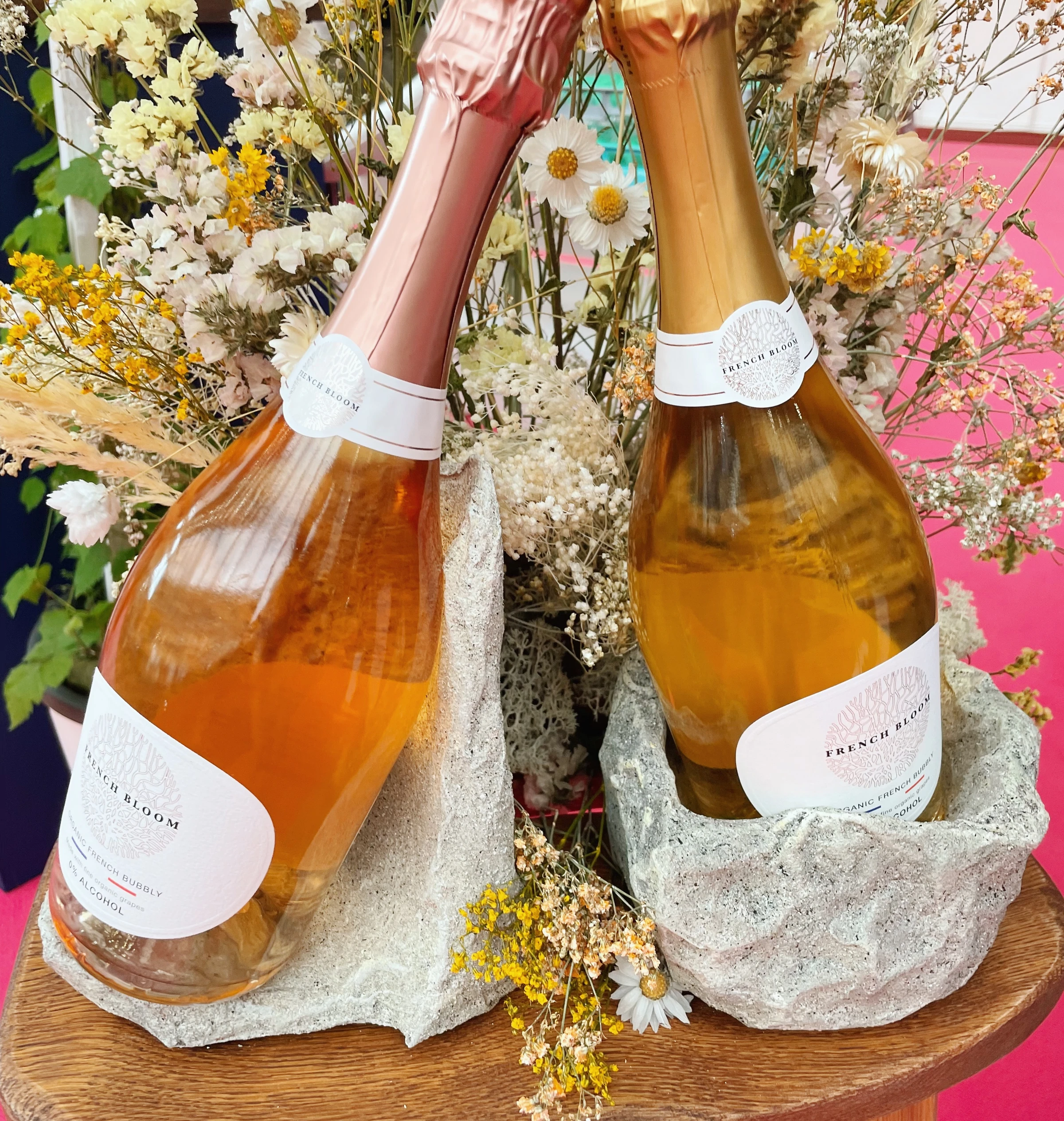 Two French Bloom alcohol-free sparkling wine bottles among dried flowers on a wooden table