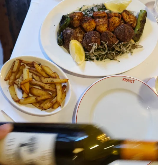 Shared plate of grilled meatballs, vegetables, chips and lemon beside a blurred wine bottle