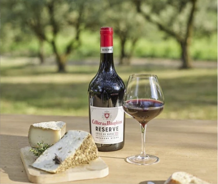 Bottle of Cellier des Dauphins Réserve red wine and a filled wine glass beside cheeses outdoors
