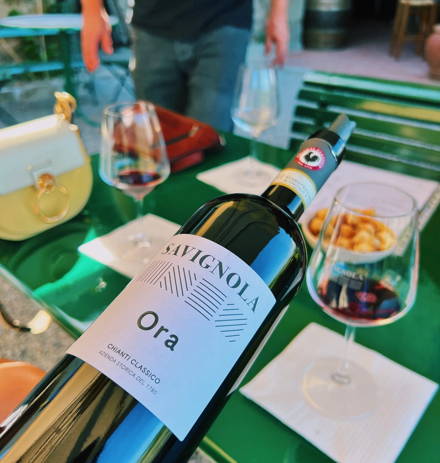 Savignola Ora wine bottle beside tasting glasses and snacks on a green table