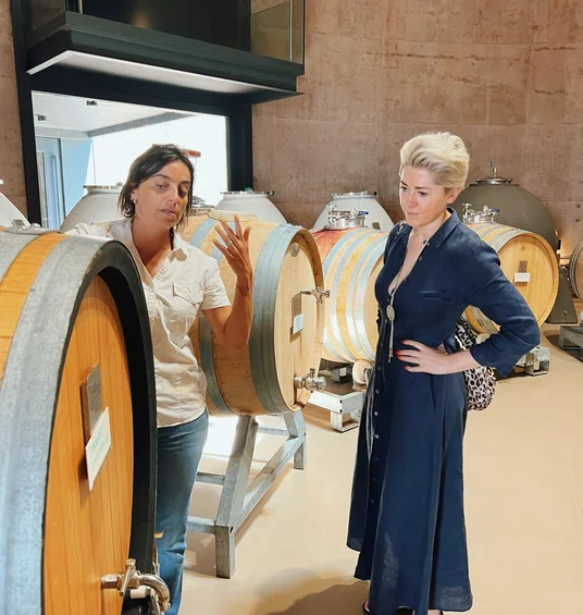 Two women discussing wine production beside oak barrels in a modern cellar