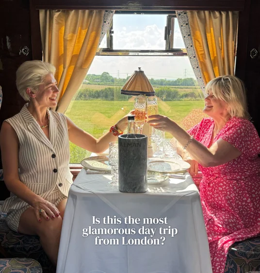 Two women toast aboard a luxury train beside a sunlit countryside view