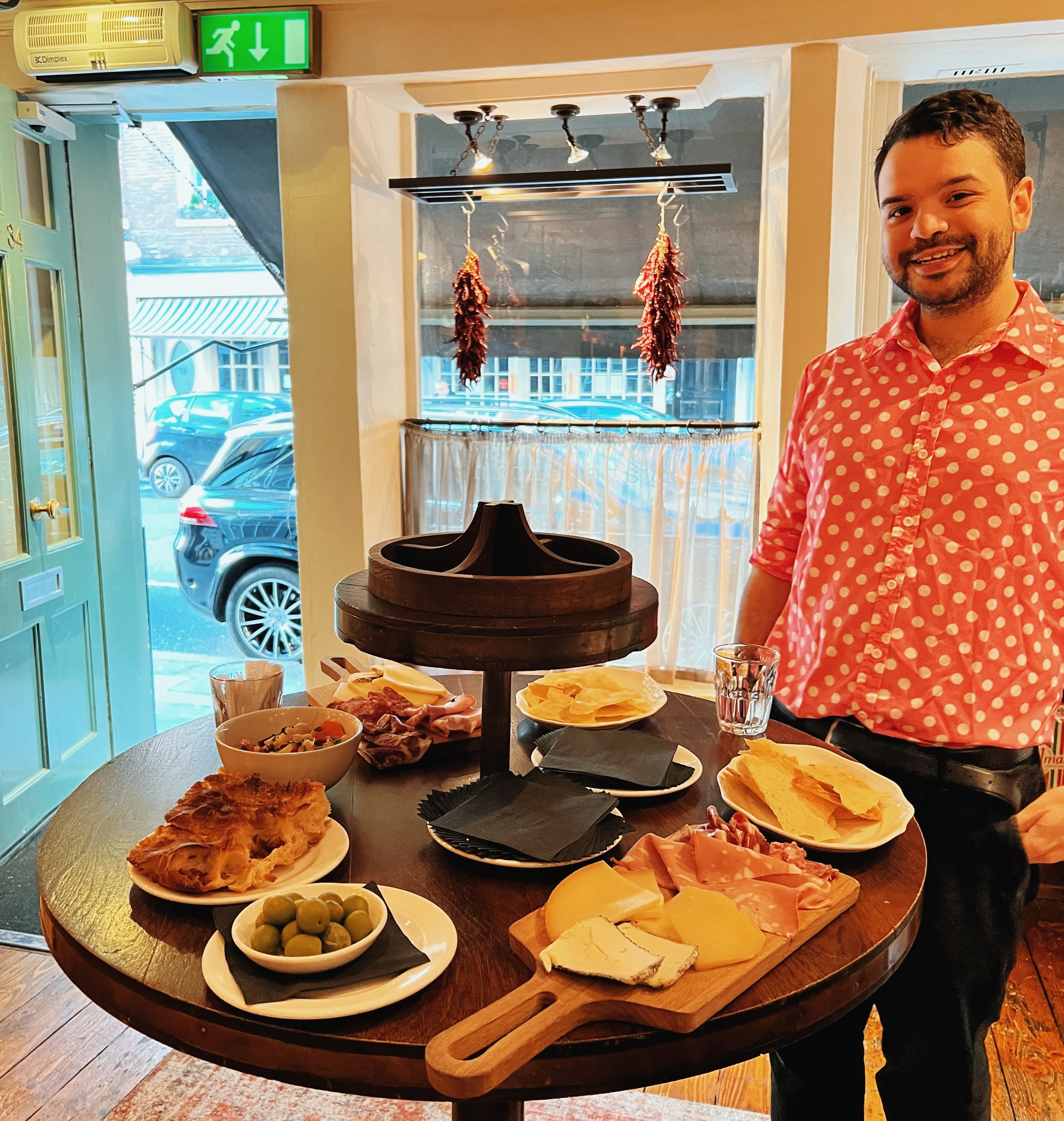 Man in a pink patterned shirt beside a table laden with cheeses, charcuterie, bread, crisps and olives