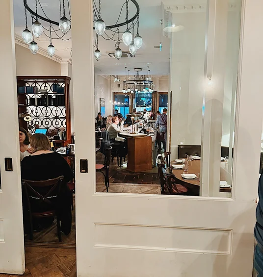 Elegant restaurant interior viewed through mirrored doors, with diners seated beneath circular pendant lights
