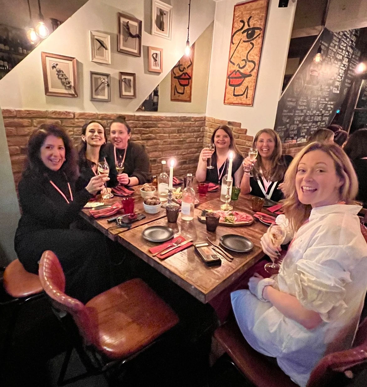Six women raise wine glasses around a candlelit restaurant table with food and wine bottles.