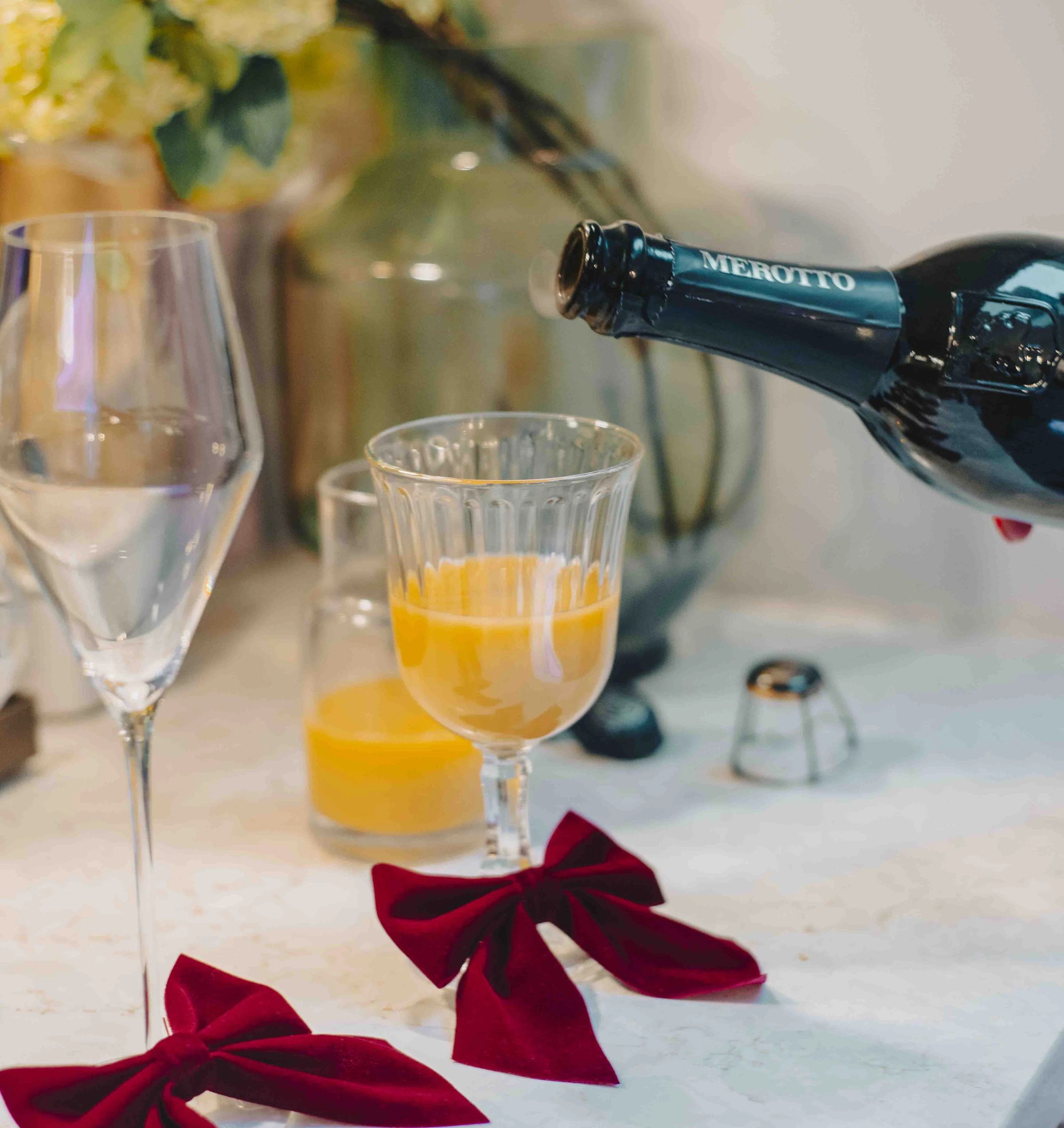 Merotto bottle pouring sparkling wine into an orange cocktail beside red velvet bows