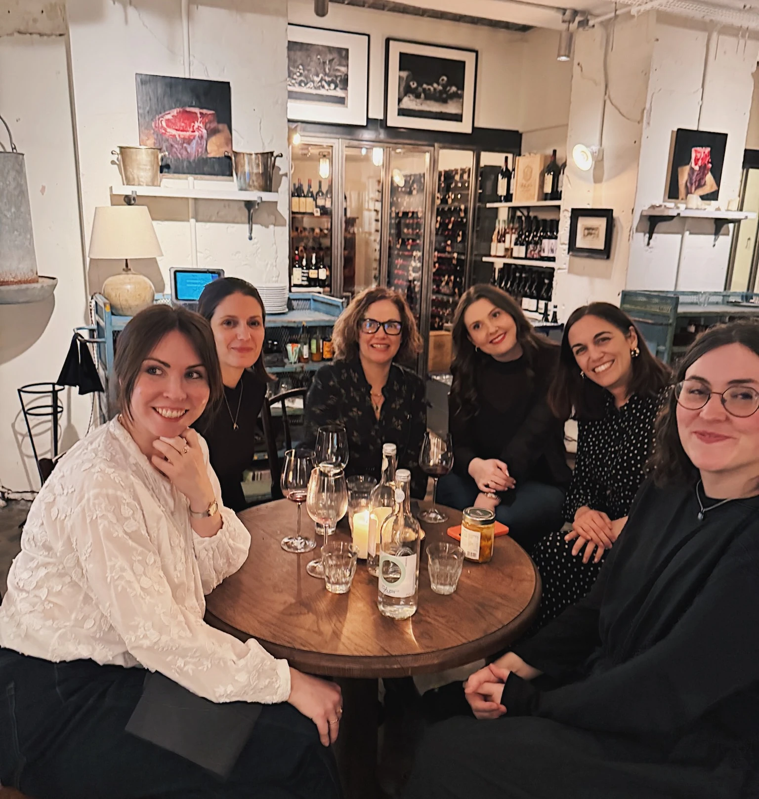 Six women seated around a table with wine glasses in a restaurant displaying bottles