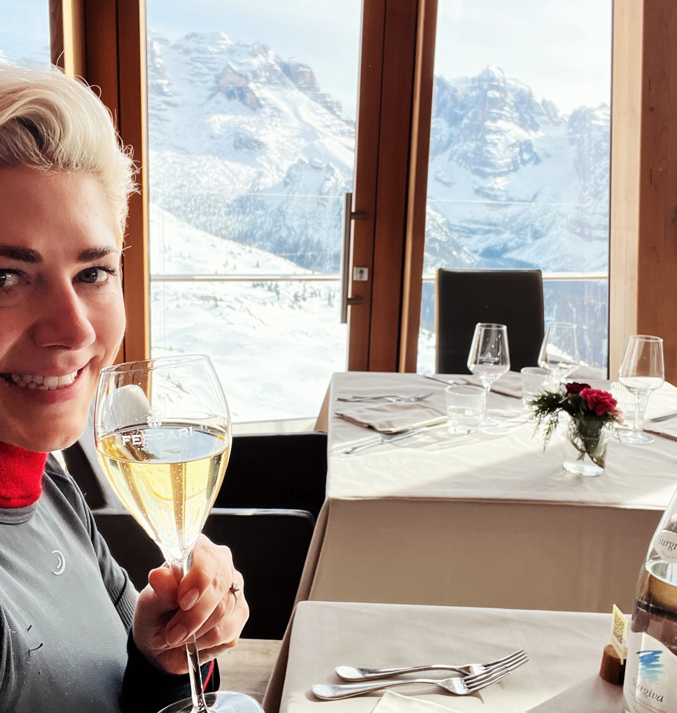 Woman holding a glass of sparkling wine beside a restaurant table with snowy mountains beyond