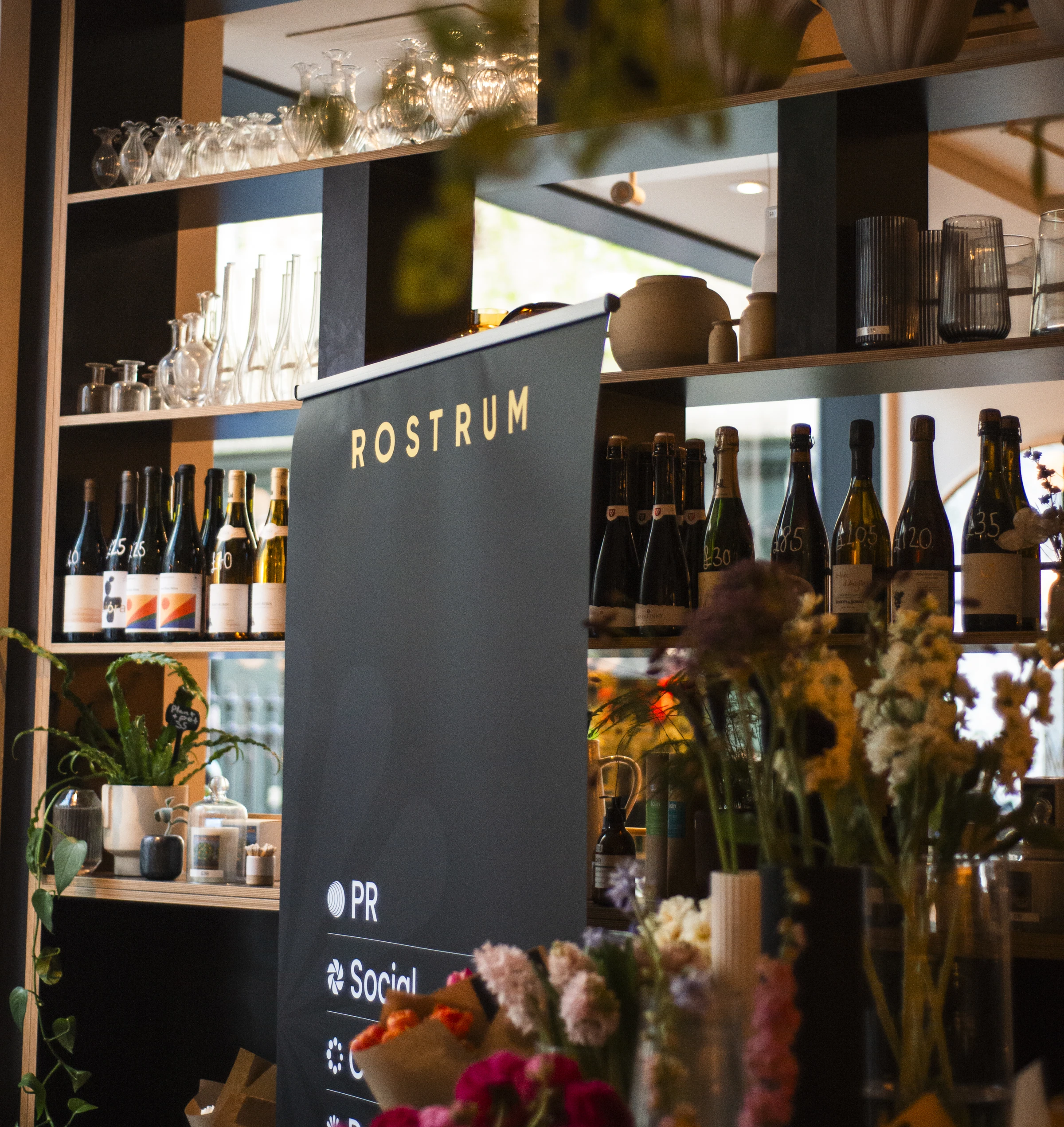 Wine bottles and glassware displayed on shelves beside a Rostrum sign and colourful flower arrangements