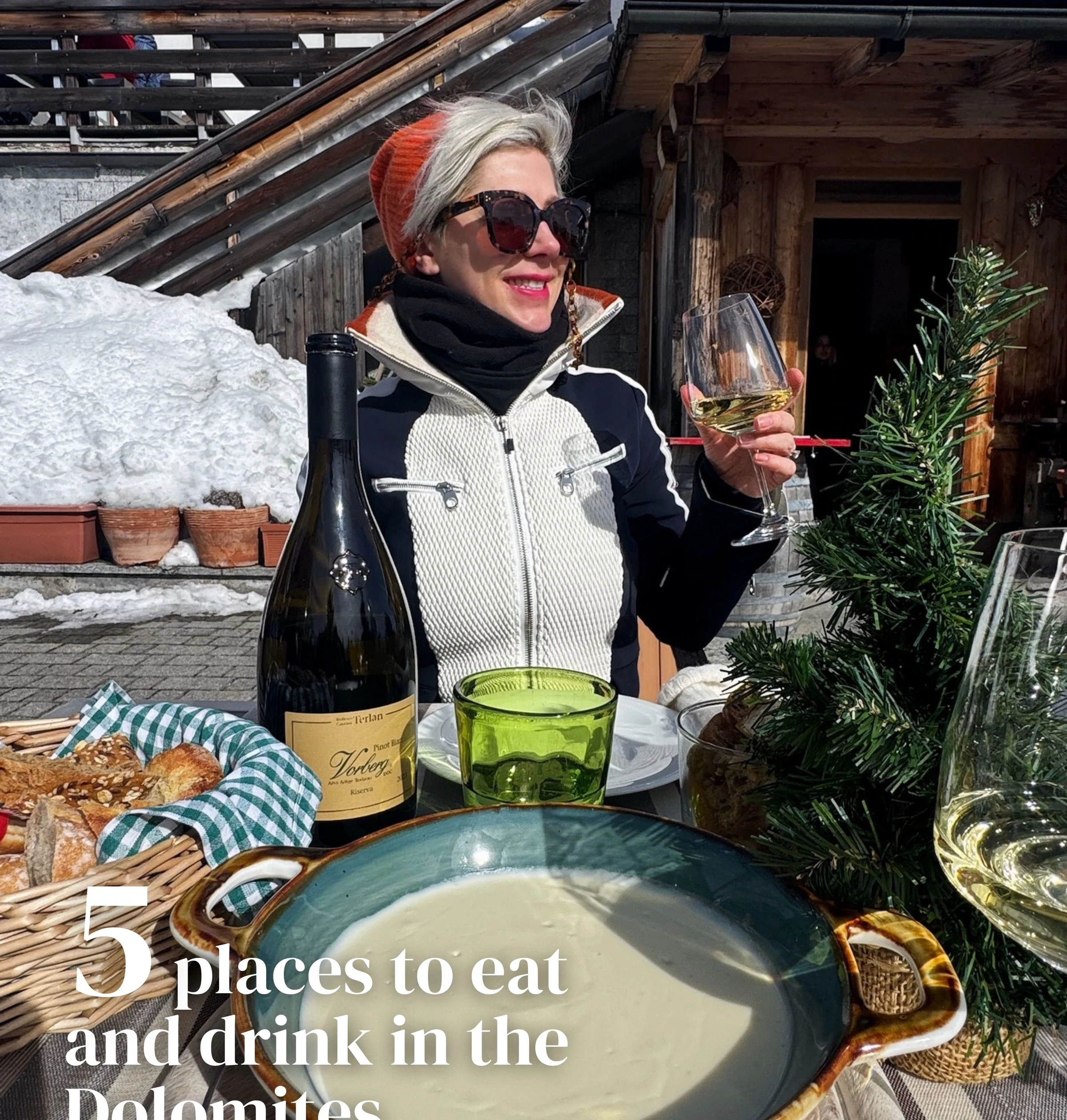 Woman holding a glass of white wine beside a bottle and alpine restaurant table in the snow
