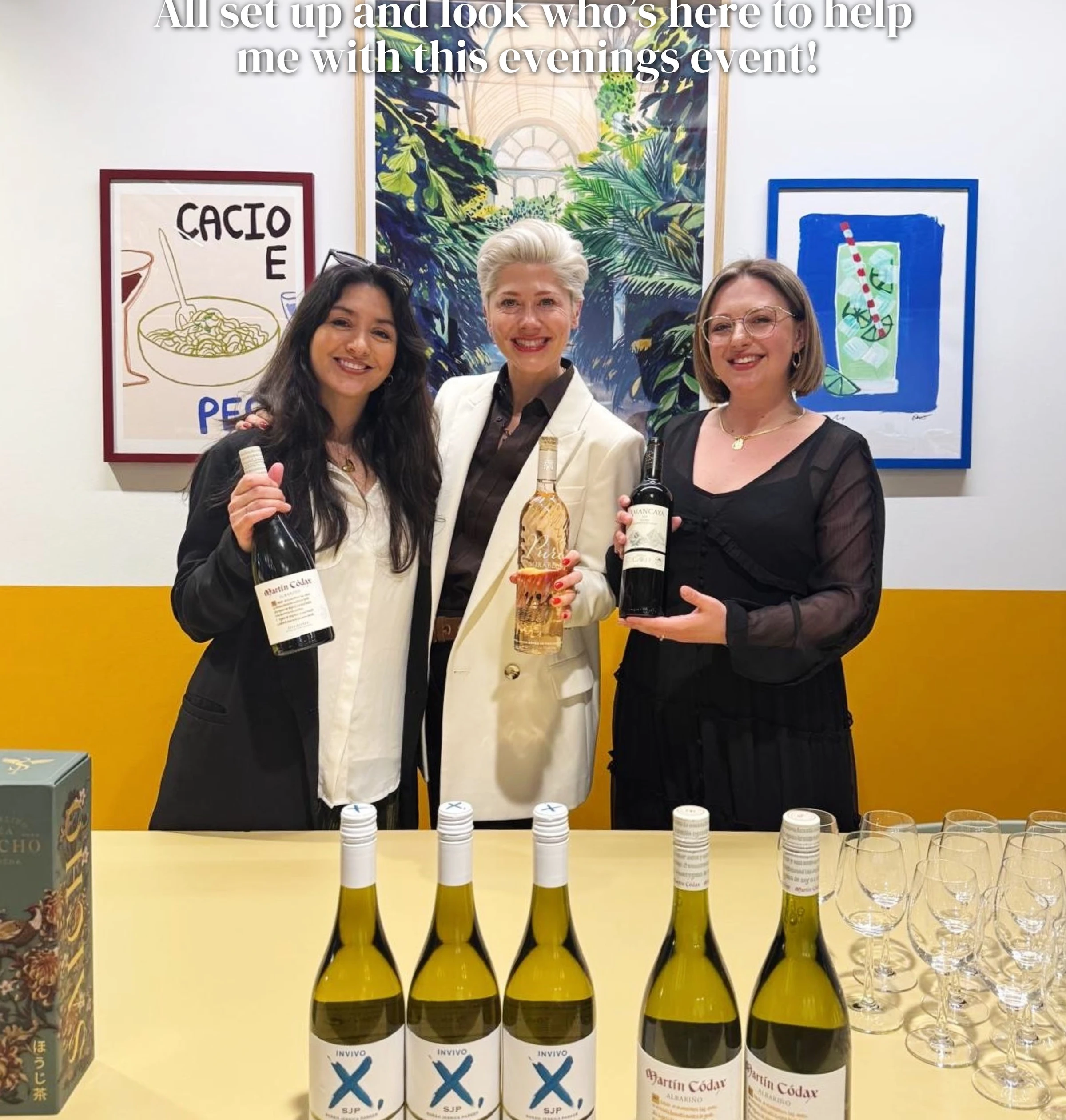 Three women pose with wine bottles beside arranged tasting glasses