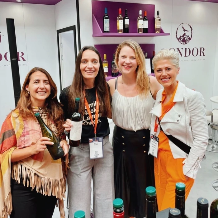 Four women smile at a wine event, with wine bottles displayed on shelves behind them.