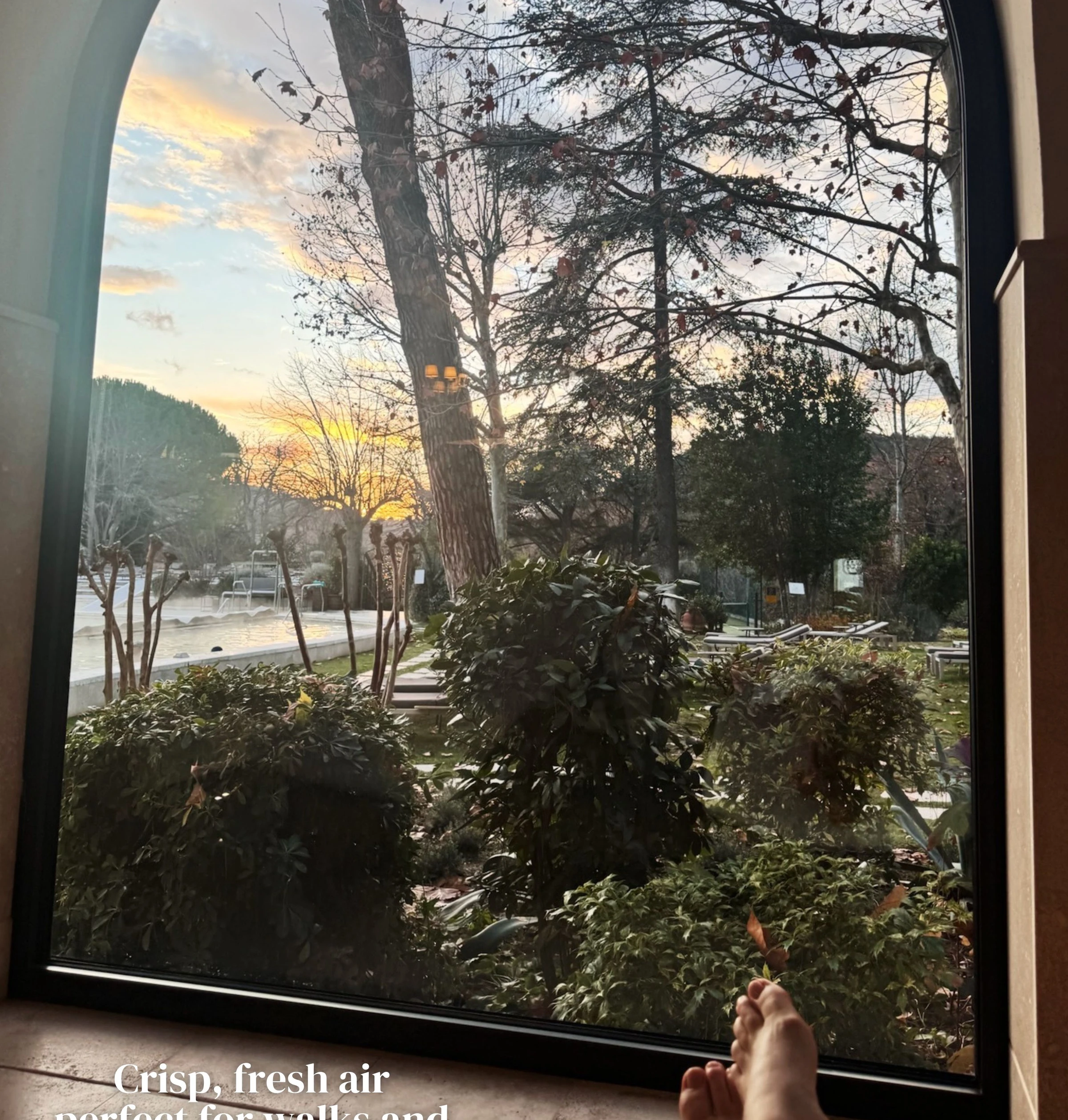 View through an arched window over a garden pool at sunset, with bare feet in the foreground