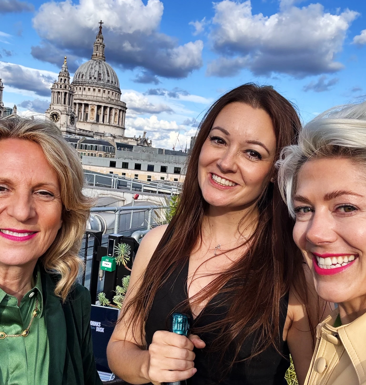 Three women pose with a wine bottle on a rooftop terrace, with St Paul's Cathedral behind them