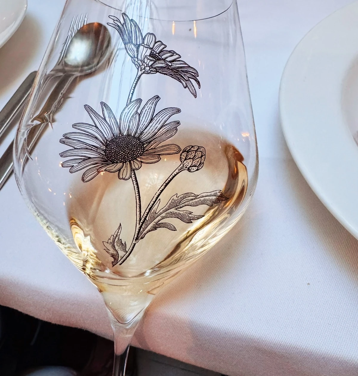 Glass of rosé wine with black floral illustrations, held over a white tablecloth beside plates and cutlery.