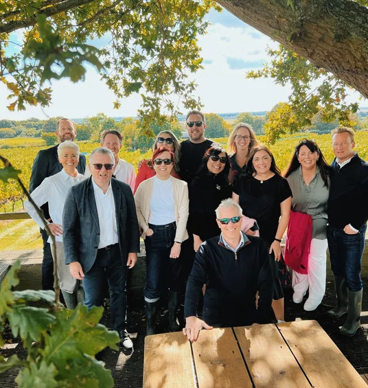 Group of visitors posing beneath a tree overlooking vineyard rows during an English vineyard visit