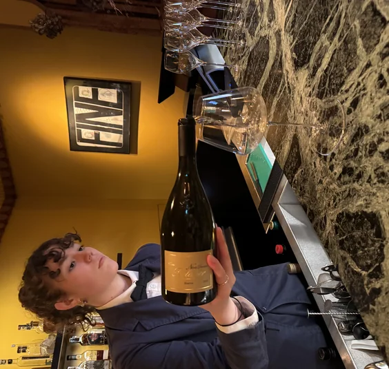 Bartender holding a bottle of wine behind a bar, with wine glasses and bottles visible