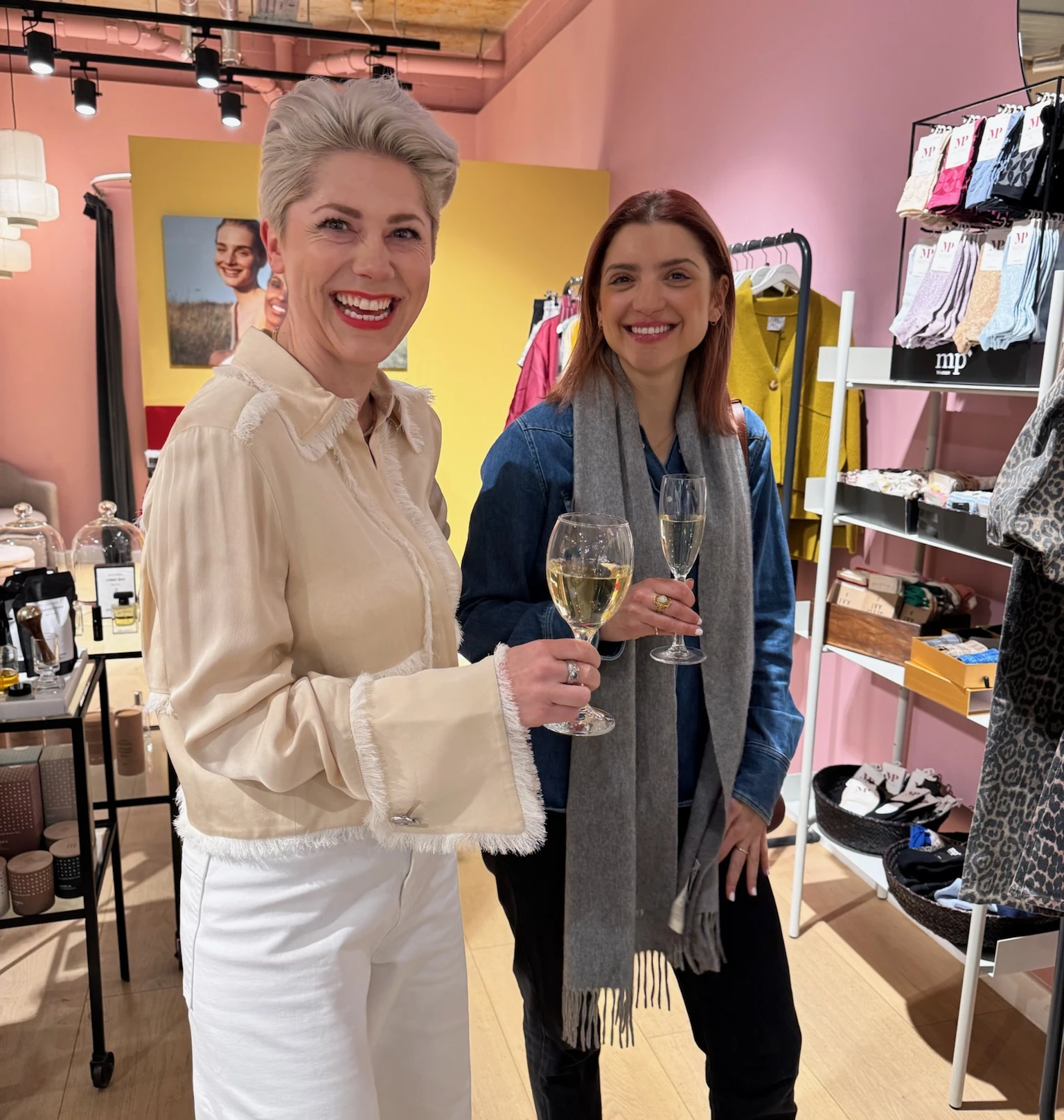 Two women smiling and holding glasses of white wine in a colourful clothing shop