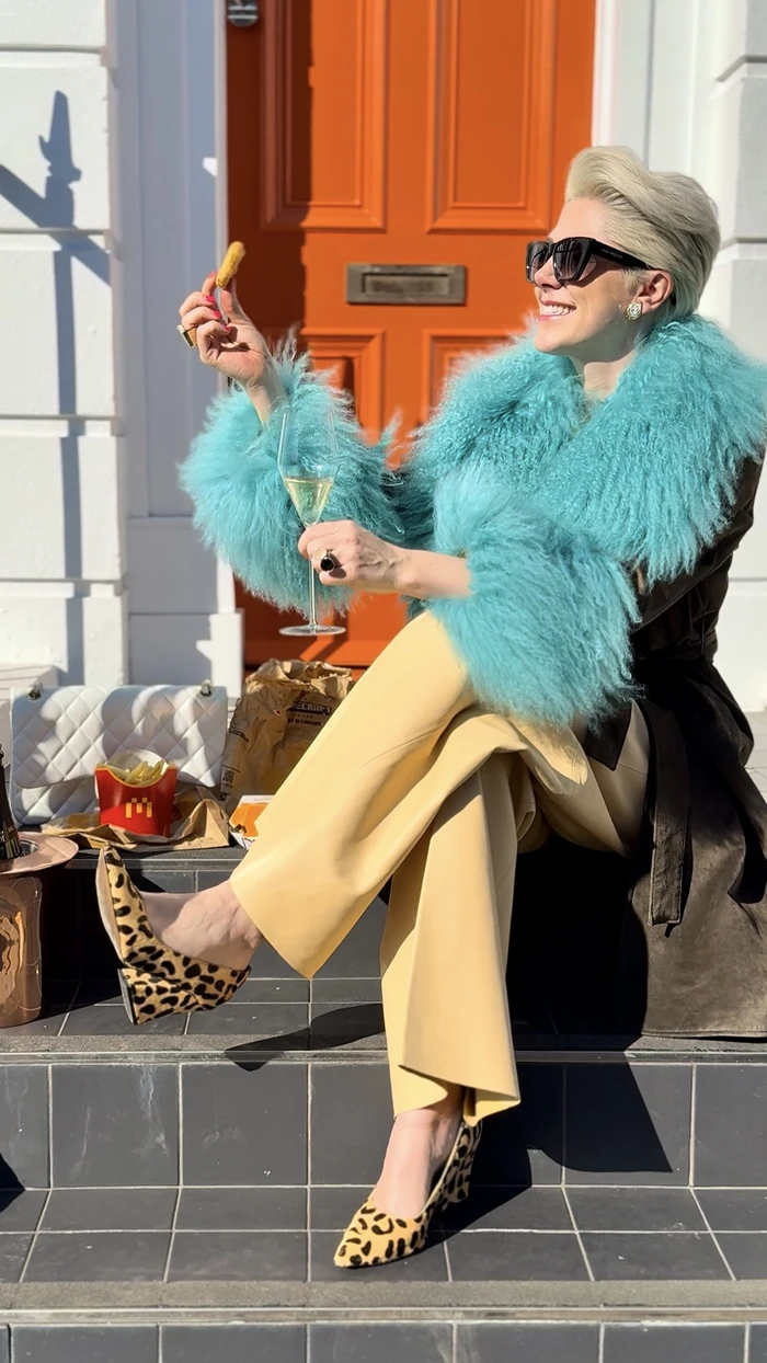 Woman in turquoise faux fur coat holding champagne and snack outdoors