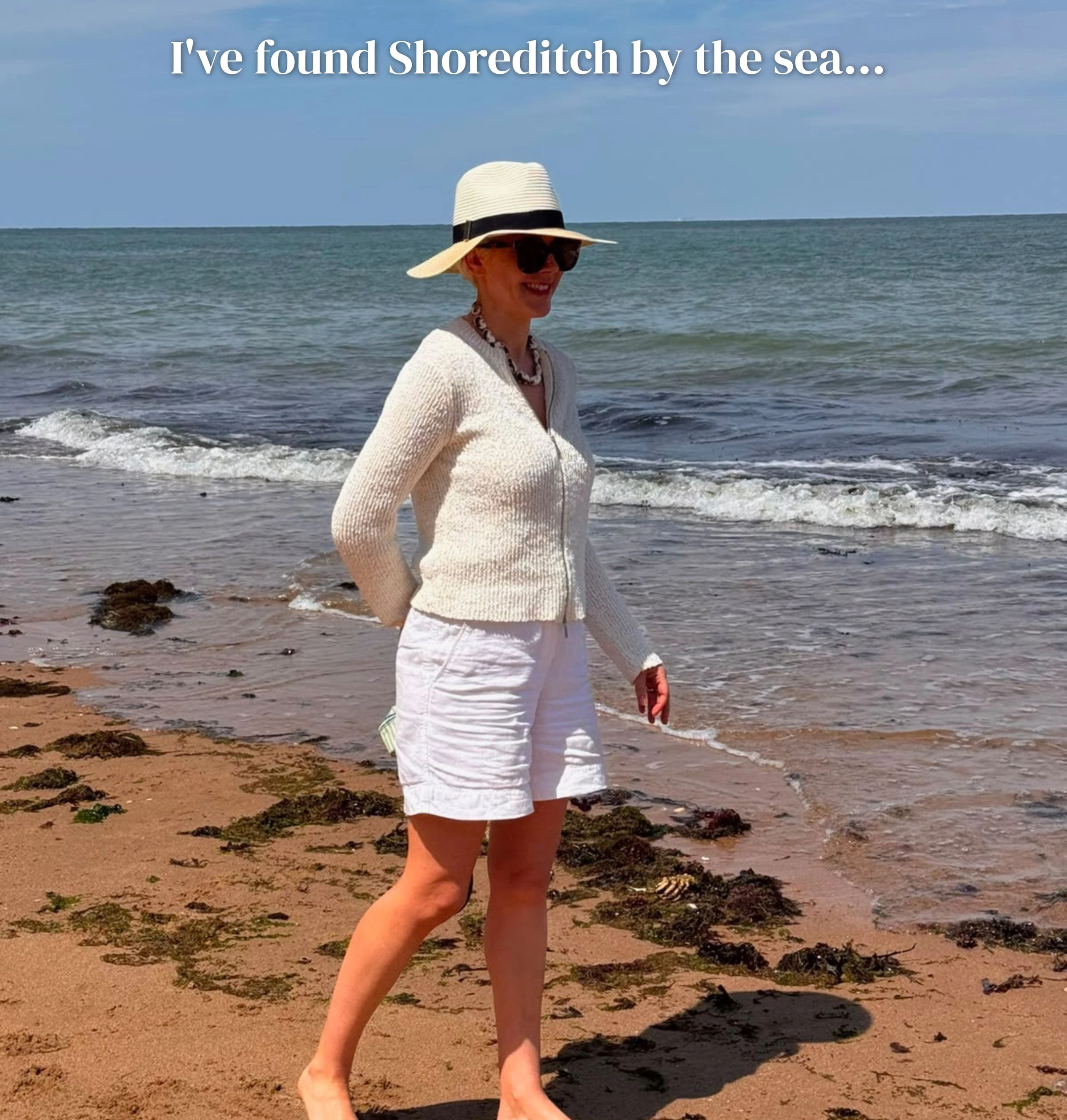 Person in a sun hat and white clothing standing barefoot on a beach beside the sea