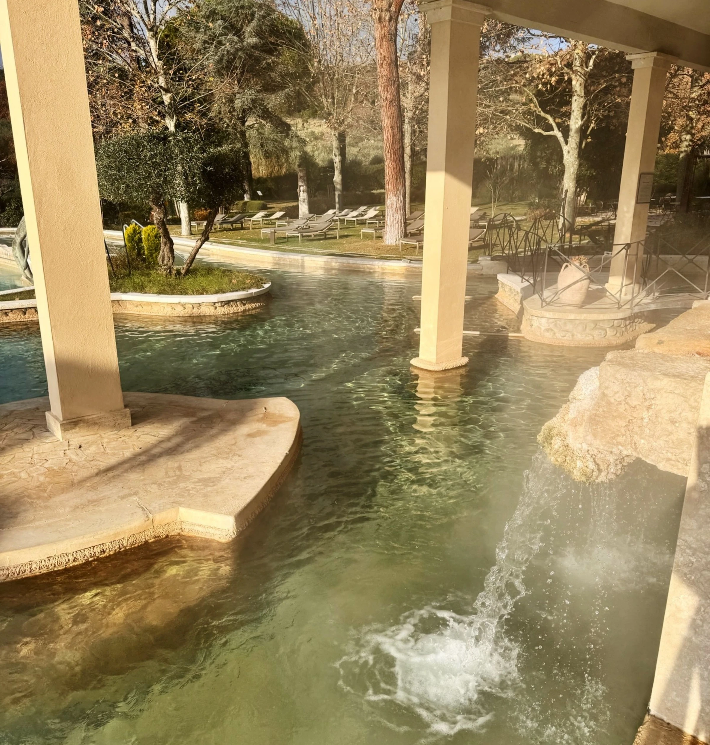 Thermal spa pool beneath a covered terrace, with landscaped gardens and loungers beyond