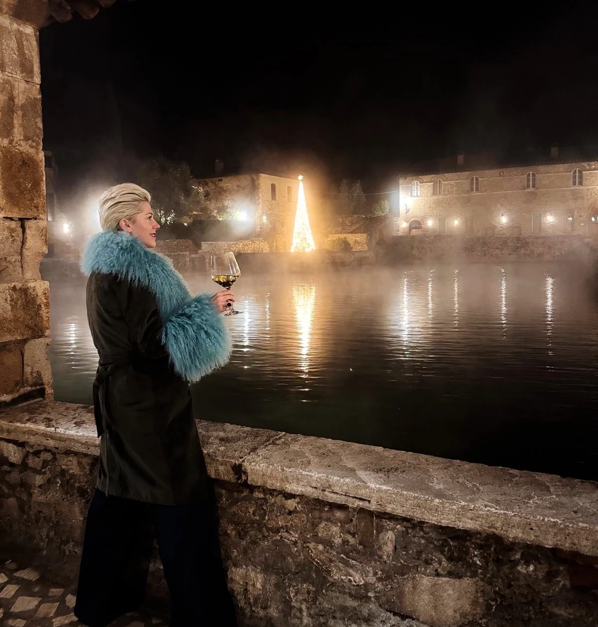 Person holding a wine glass beside a misty pool with a lit Christmas tree at night
