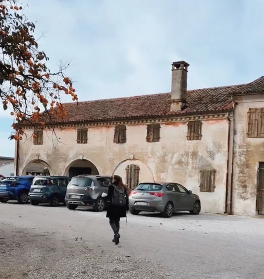 Person walking towards a weathered Italian building with arched openings, shuttered windows and parked cars