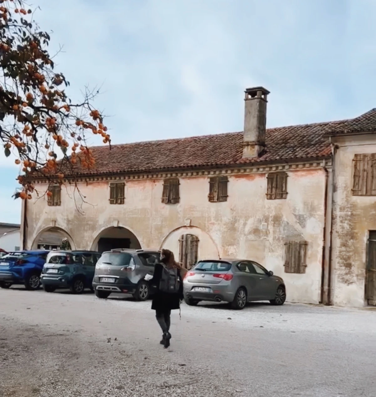 Person walking towards a weathered Italian building with arched openings, shuttered windows and parked cars