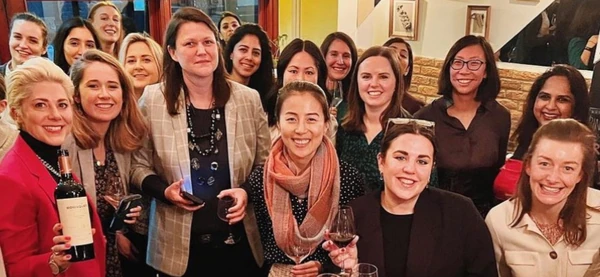 Group of people smiling indoors, with two women holding wine glasses and one holding a bottle