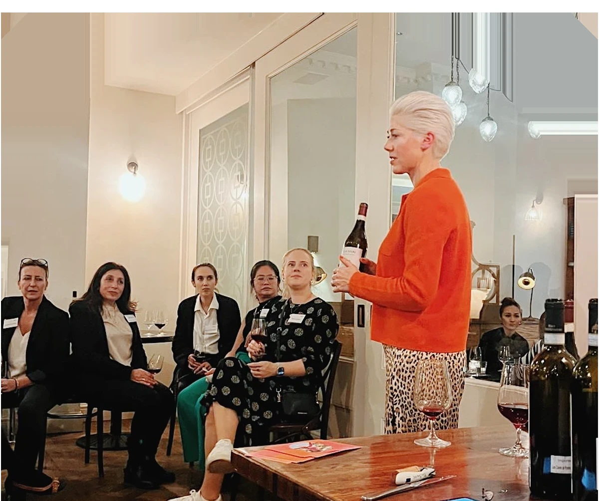 Person presenting a wine bottle to seated attendees during a wine tasting event