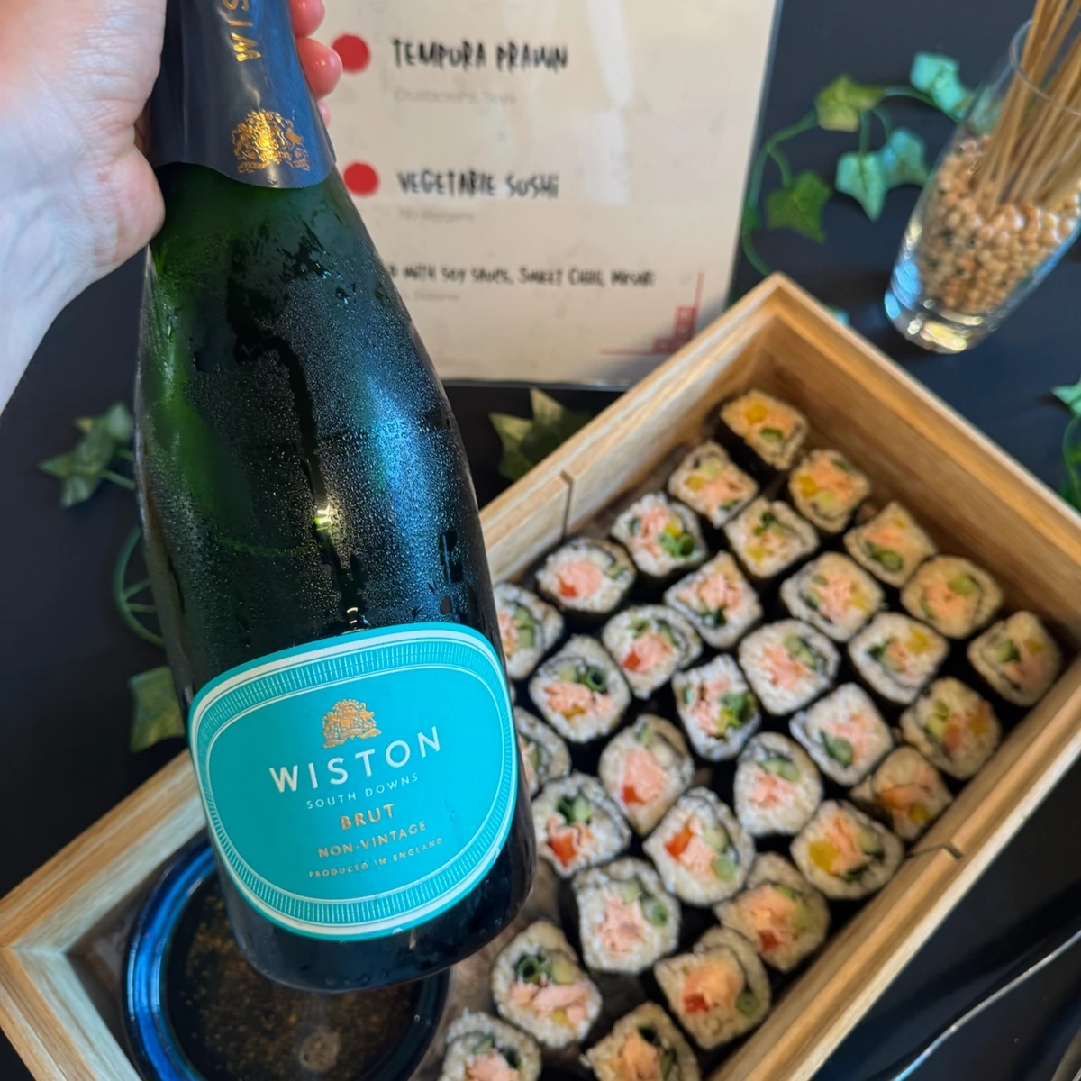 Hand holding a bottle of Wiston South Downs Brut beside a box of sushi rolls