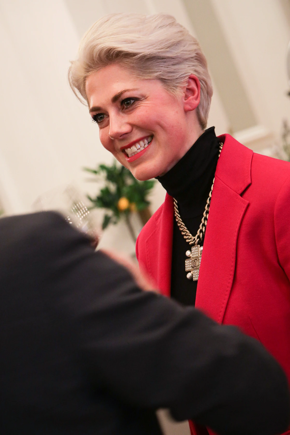 Smiling woman in a red blazer and black top talking to someone indoors