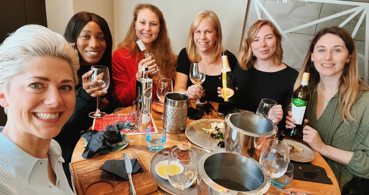 Six women smiling around a table with wine bottles, glasses and food