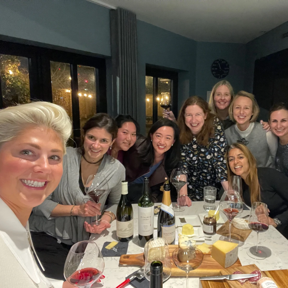 Group of women smiling around a table with wine bottles, glasses and cheese boards