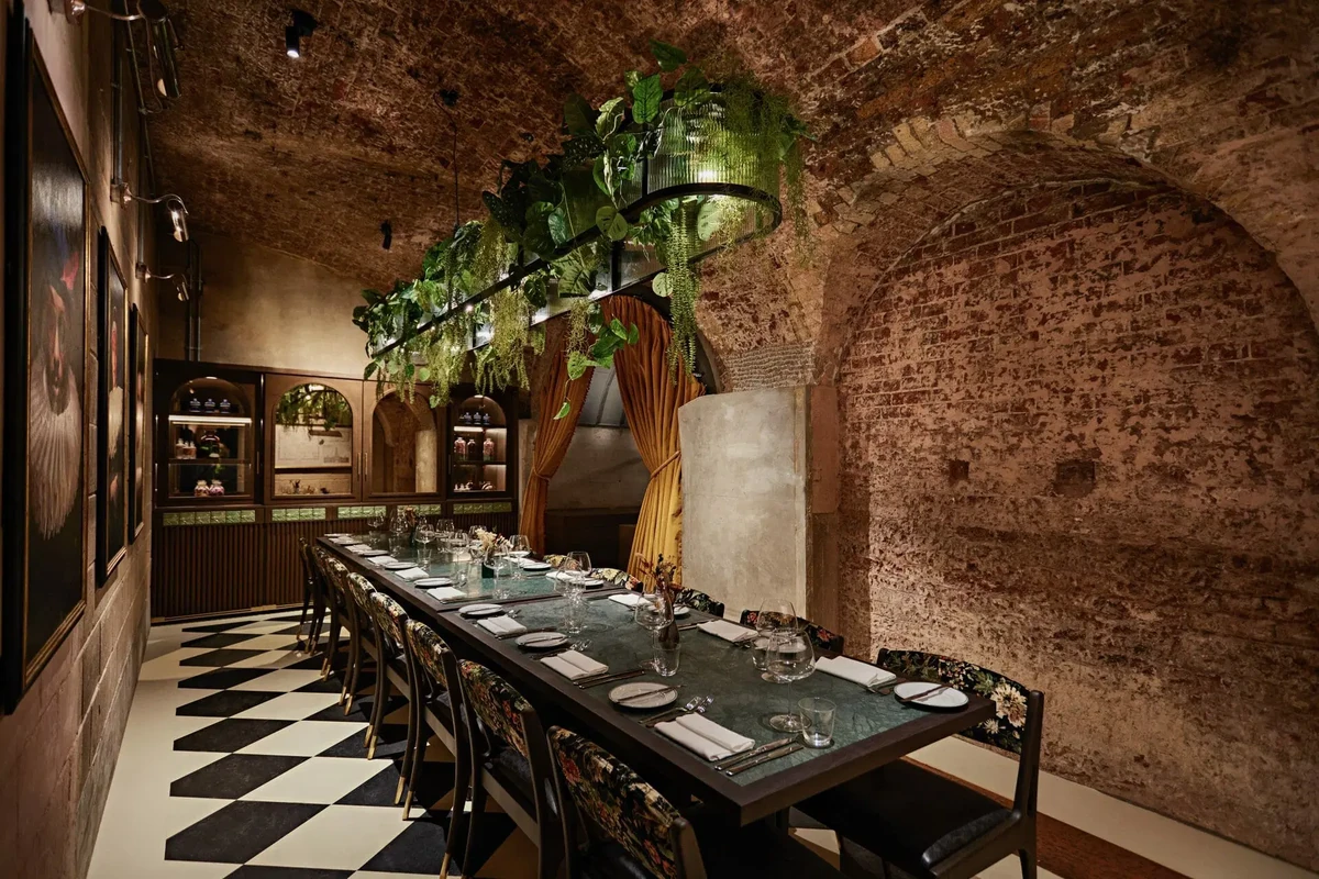 Long dining table set with glassware beneath hanging plants in a brick-vaulted restaurant